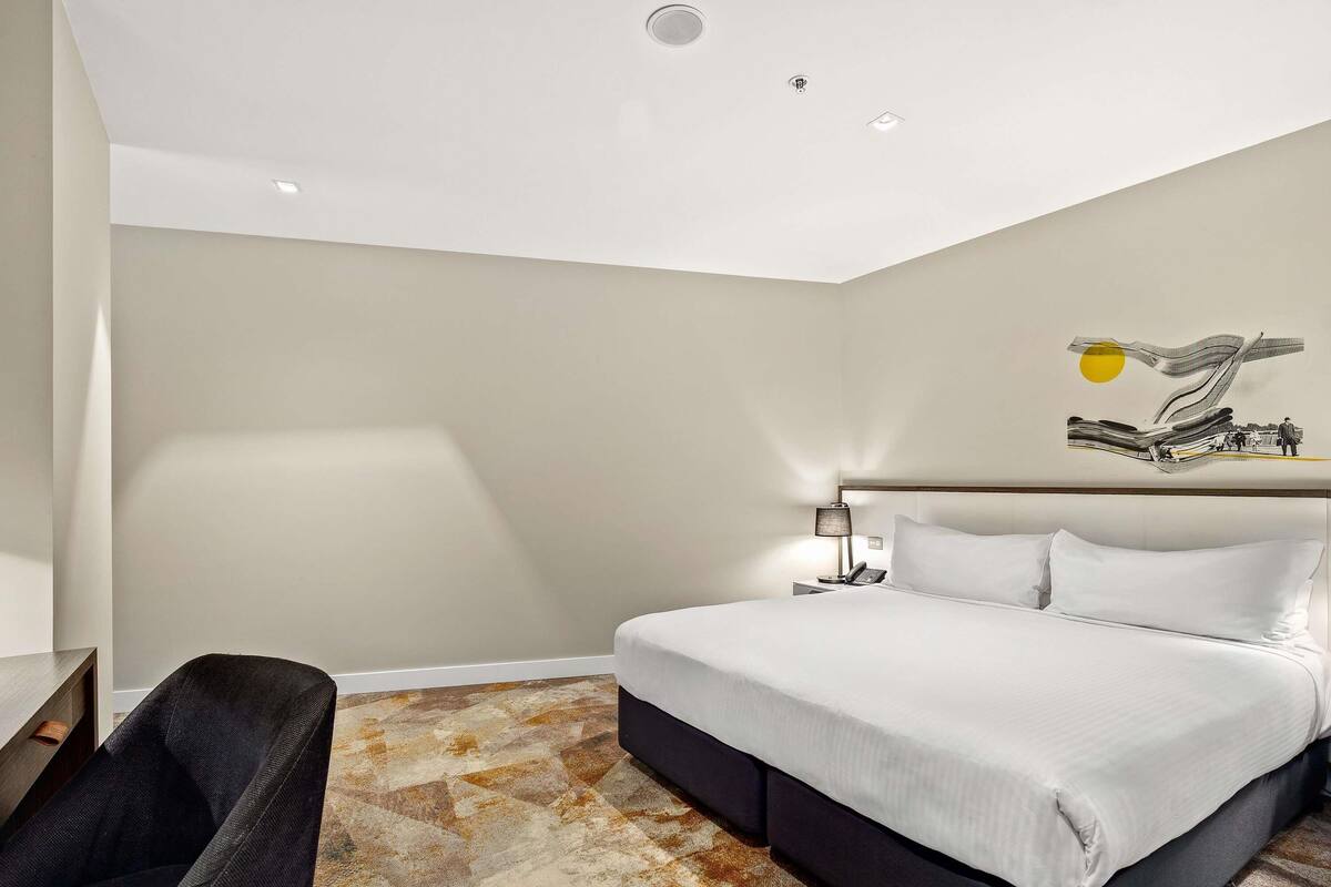 Quarto de hotel com cama, cadeira, luminária e uma obra de arte na parede.
