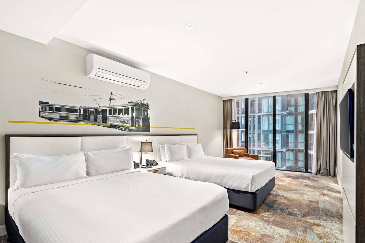 Quarto de hotel com duas camas, uma janela grande com cortinas, ar condicionado fixado na parede e televisão na parede.