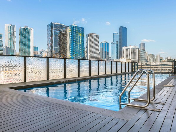 A rooftop pool with a city skyline view, metal railings, and a wooden deck.