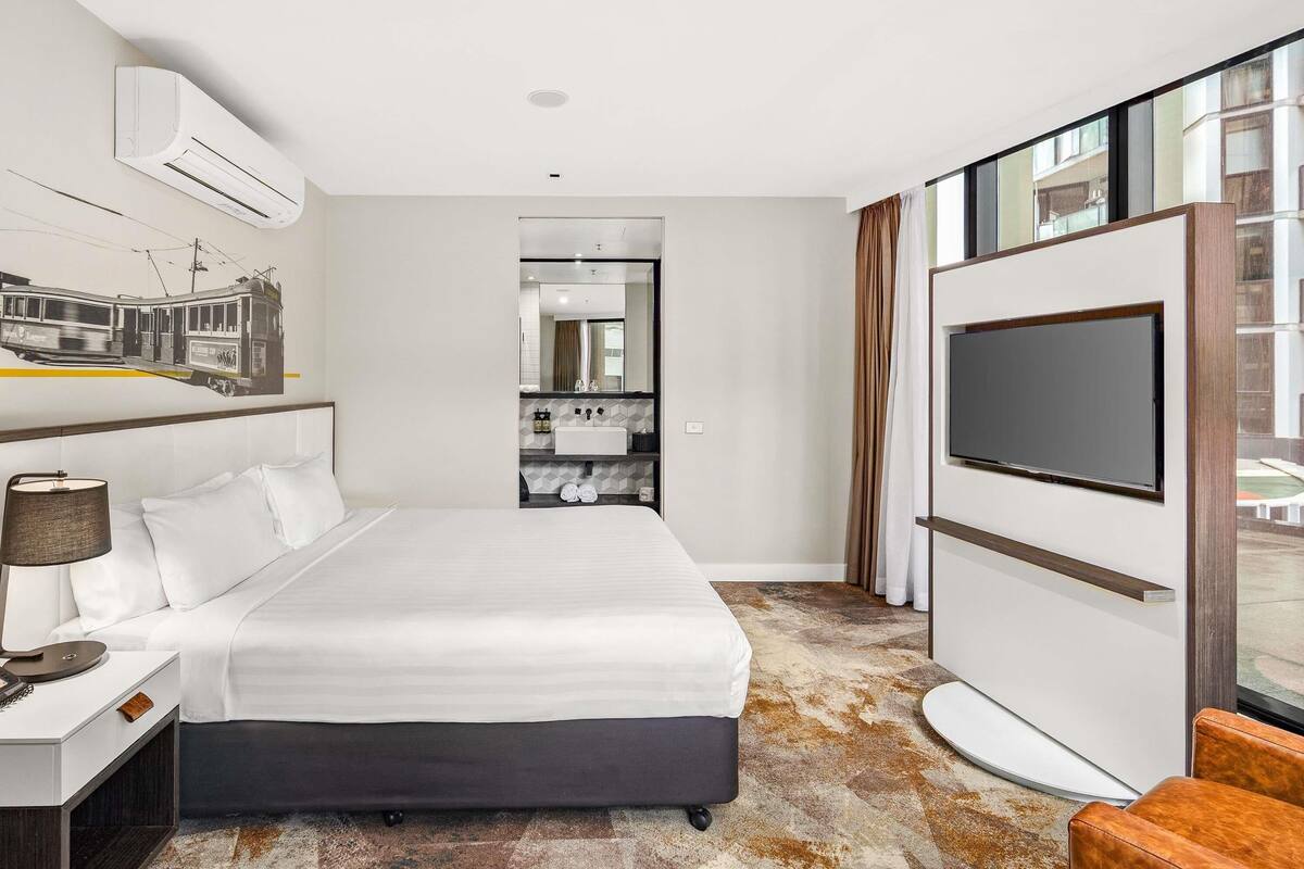 Quarto de hotel com cama, televisão, espelho e banheiro visível por uma porta aberta.