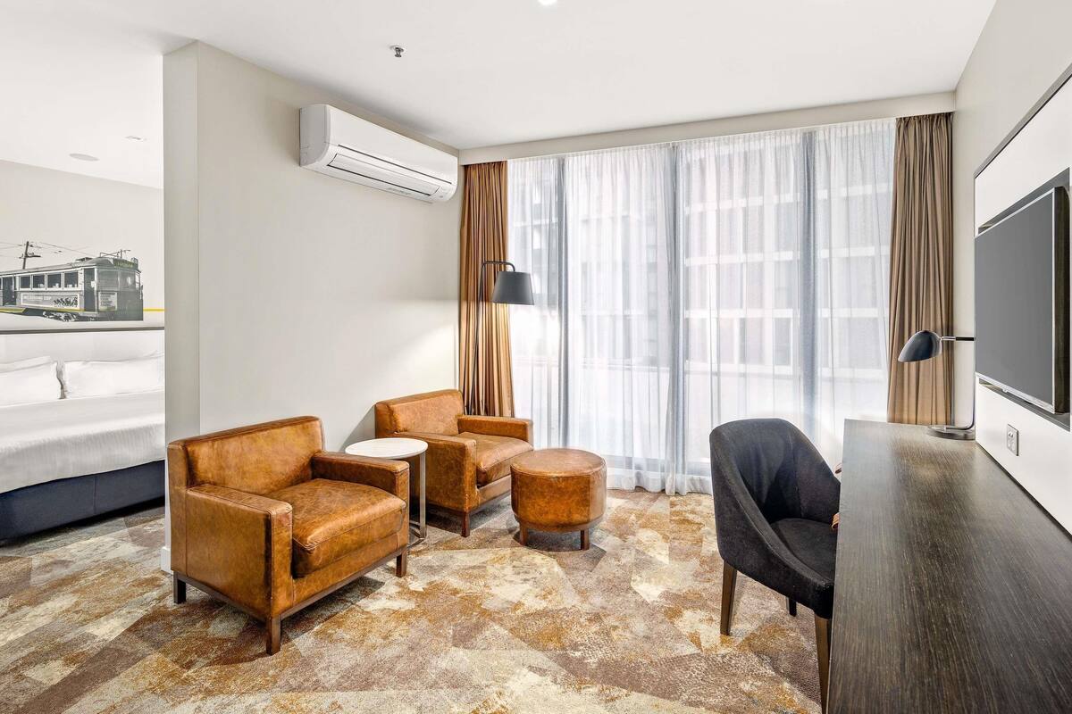 Quarto de hotel com cama, duas poltronas de couro, uma mesa redonda pequena, escrivaninha com cadeira, televisão, aparelho de ar condicionado e uma janela grande com cortinas.