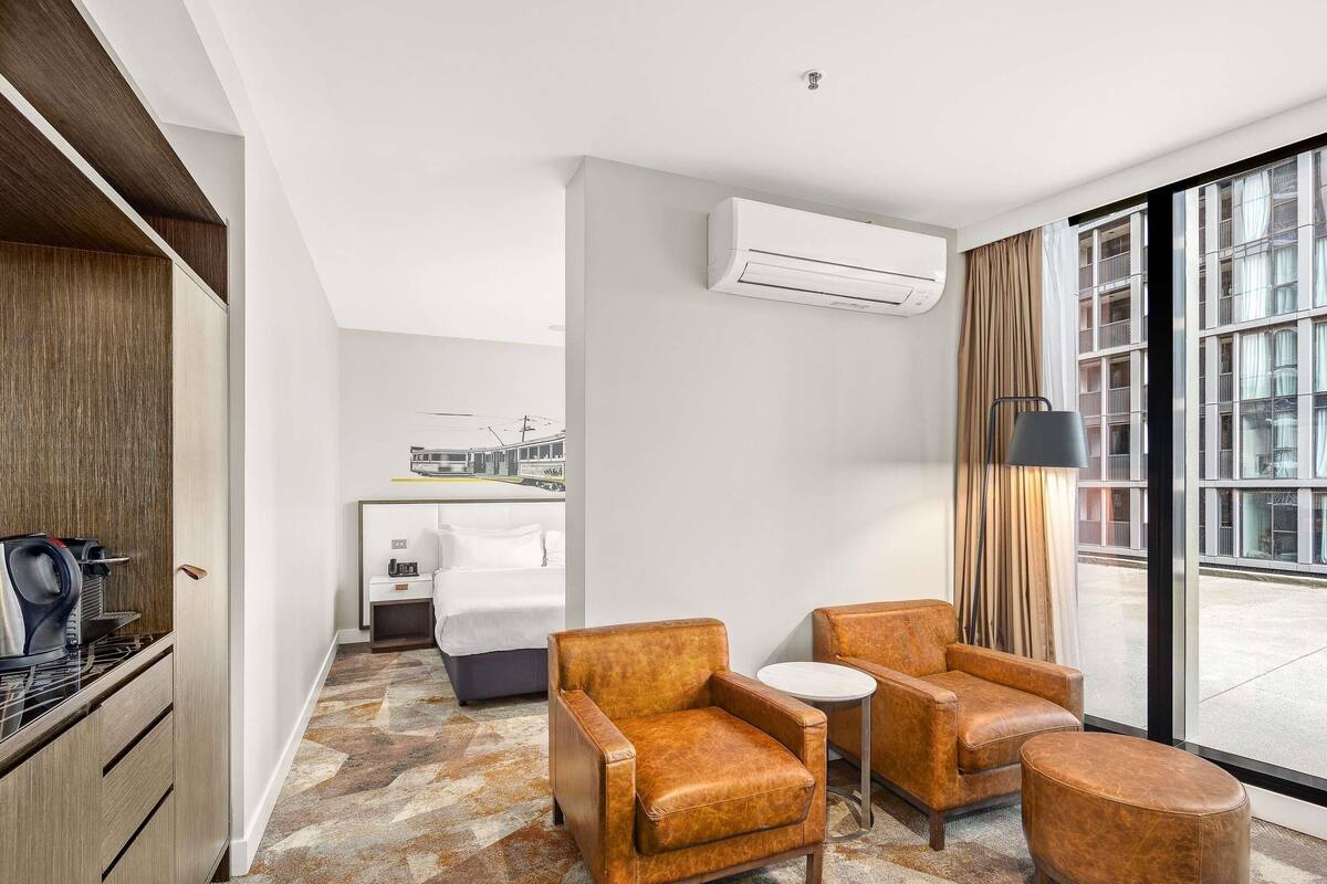 Quarto de hotel com cama, secretária com máquina de café, duas poltronas de couro, uma mesinha e uma janela grande com cortinas.