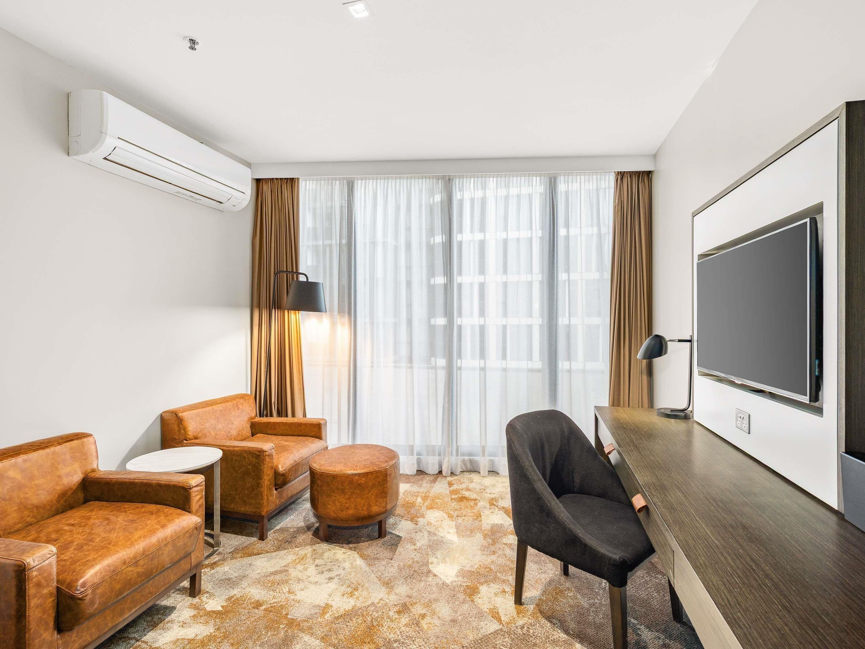 Une chambre d’hôtel équipée d’un canapé, d’un fauteuil, d’un pouf, d’un bureau avec une chaise, d’une grande télévision et d’un appareil de climatisation.