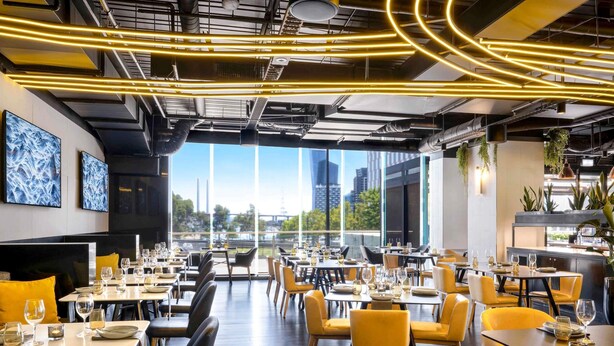 A modern restaurant with high ceilings, exposed ductwork, and a view of the cityscape. Tables are set with wine glasses and plates, and yellow chairs are arranged around them. - Peppers Southern Cross Melbourne (Docklands)
