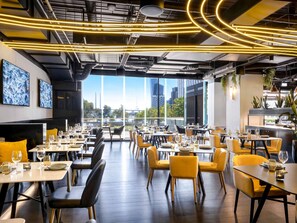Um restaurante moderno com pé-direito alto, dutos aparentes e vista para a paisagem urbana. Mesas montadas com copos de vinho e pratos, e cadeiras amarelas dispostas ao redor.