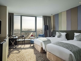A hotel room with a large window, a desk, two beds, and a view of the cityscape.