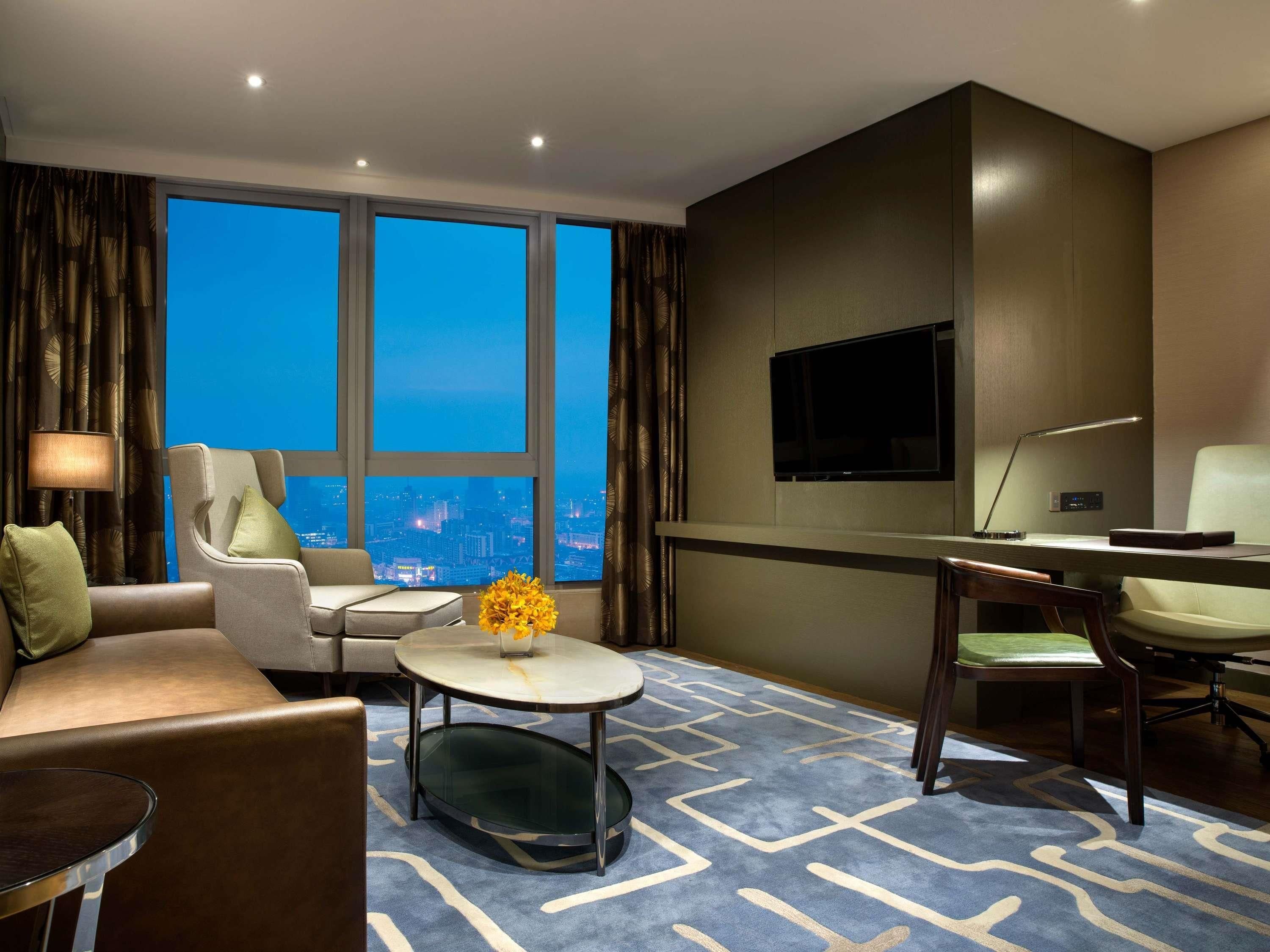 Une chambre d’hôtel équipée d’un canapé, d’un fauteuil, d’une table basse ronde, d’une télévision, d’un bureau et d’une grande fenêtre donnant sur la ville.