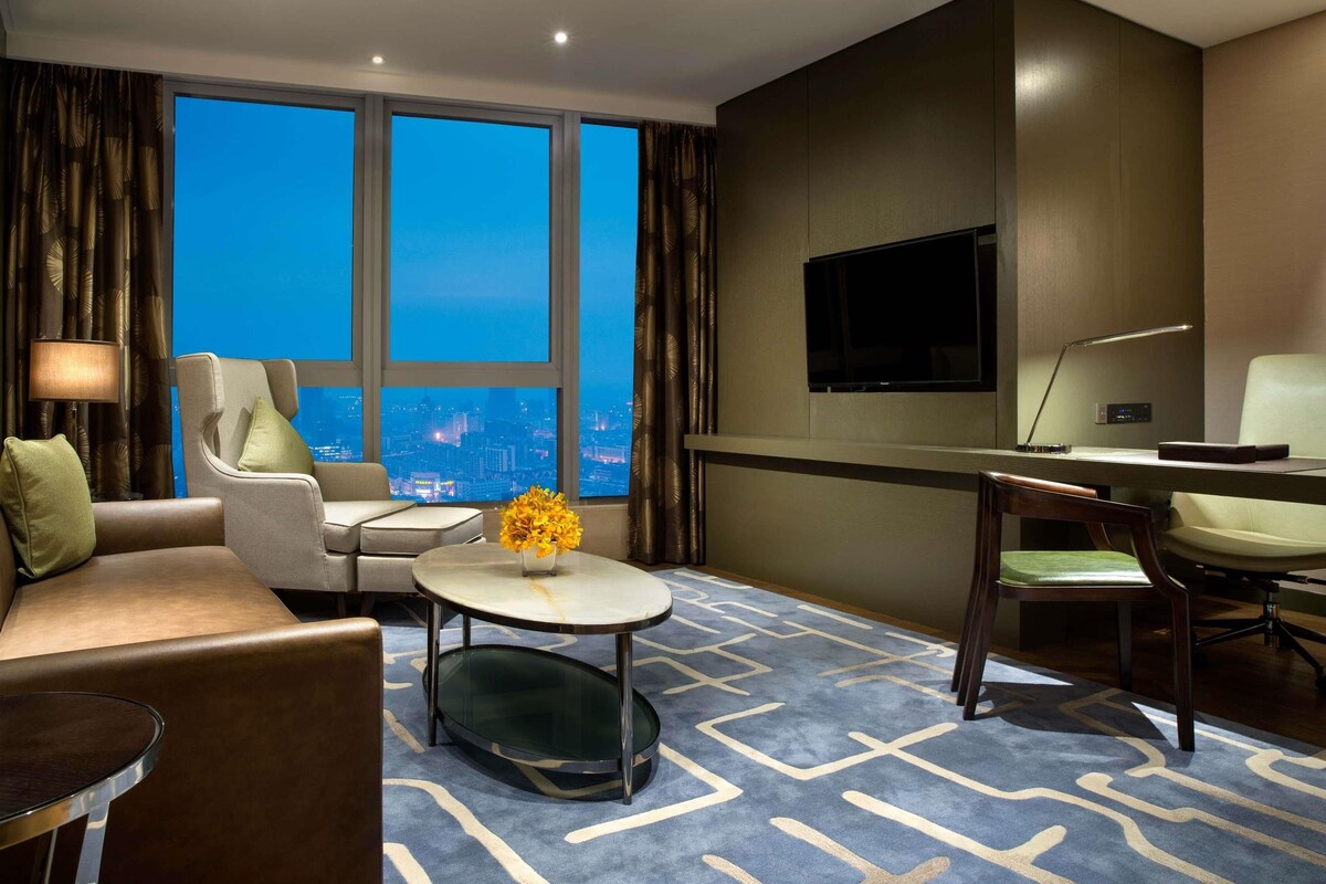 A hotel room with a sofa, armchair, round coffee table, TV, desk, and a large window with a city view.