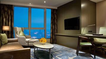 Une chambre d’hôtel équipée d’un canapé, d’un fauteuil, d’une table basse ronde, d’une télévision, d’un bureau et d’une grande fenêtre donnant sur la ville.