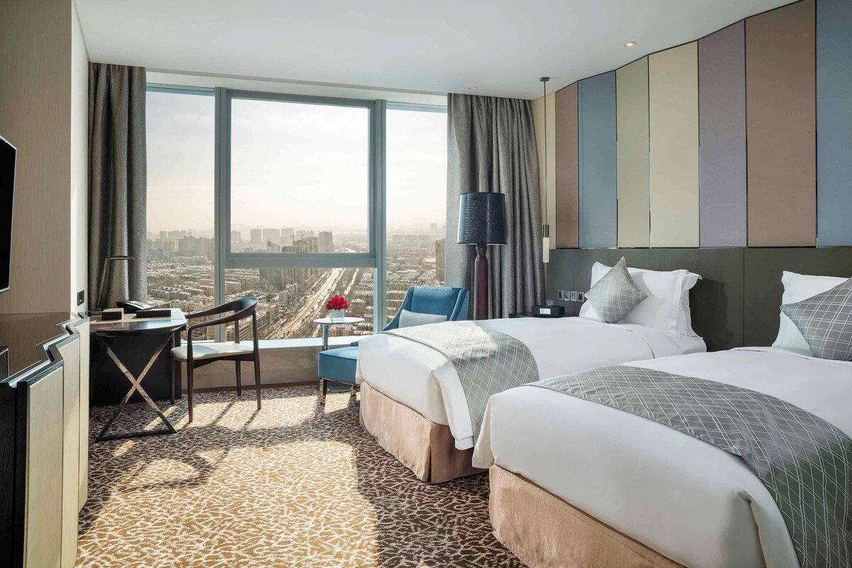 Habitación de hotel con un ventanal grande, un escritorio, dos camas y vistas a la ciudad.