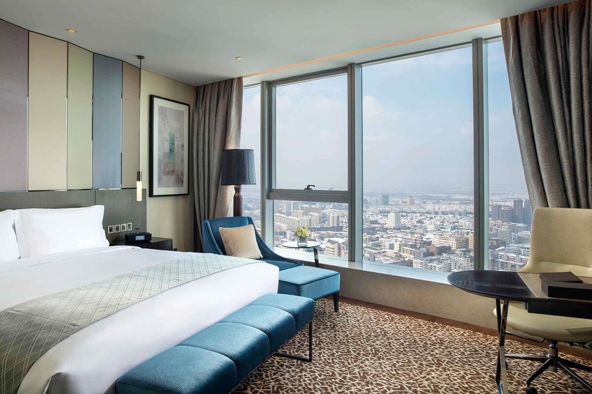 Une chambre d’hôtel avec un grand lit, un bureau, une chaise et une vue sur le paysage urbain, grâce à de grandes fenêtres.