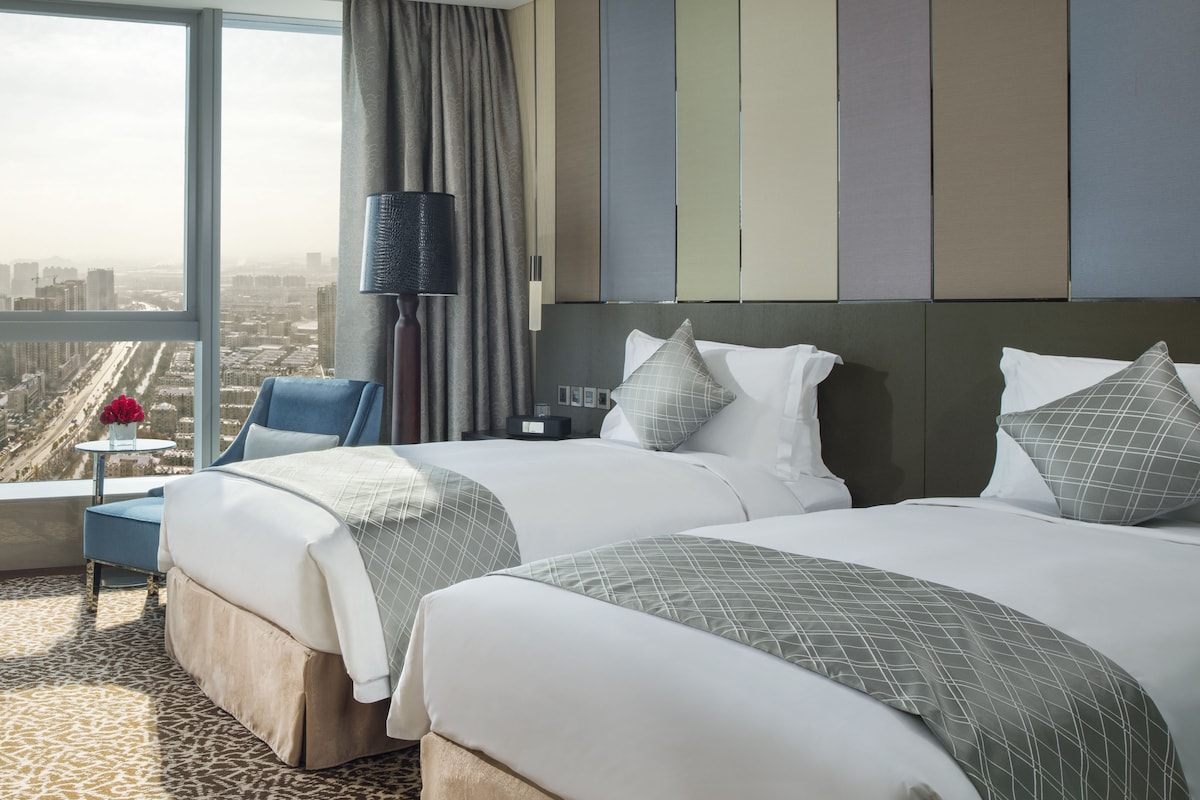 Een hotelkamer met twee bedden, uitzicht op de stad, een lamp en een klein tafeltje met een bloevas.