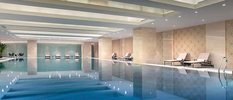 A modern indoor swimming pool with clear water, lounge chairs, and recessed lighting.