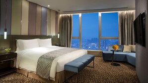 Ein modernes Hotelzimmer mit einem großen Bett, einer Sitzecke, einem kleinen Tisch und Blick auf die Stadt durch große Fenster.