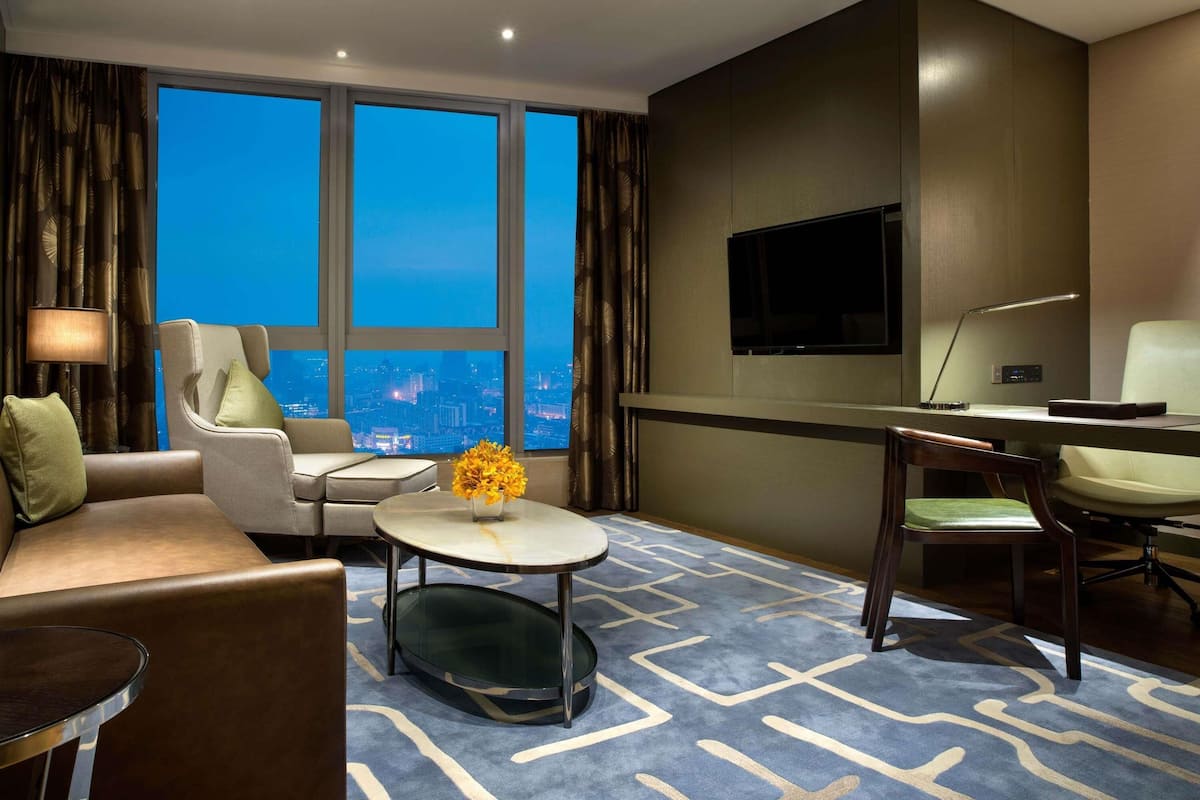Habitación de hotel con sofá, sillón, mesa de centro redonda, televisor, escritorio y un amplio ventanal con vistas a la ciudad.