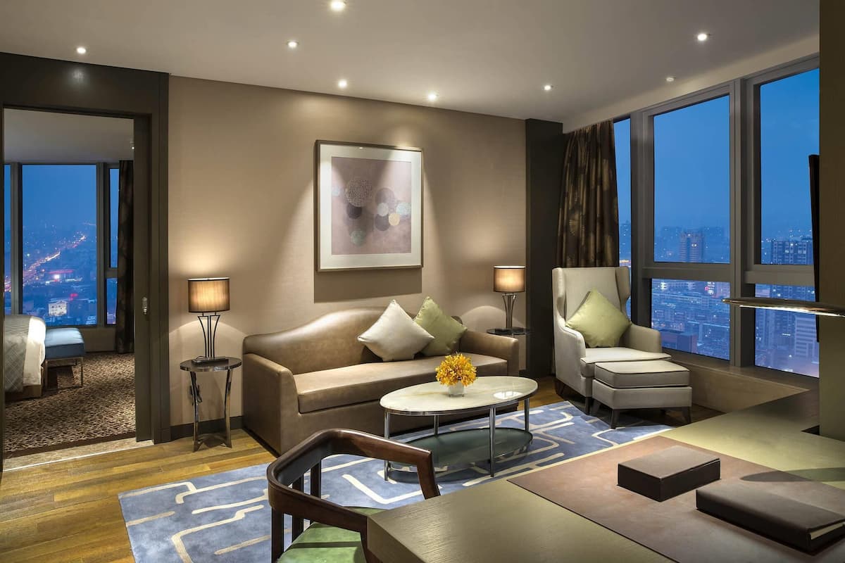 Una habitación de hotel moderna con un ventanal grande que ofrece vistas a la ciudad, un sofá, una silla, una mesita de centro y una mesita auxiliar con lámpara.