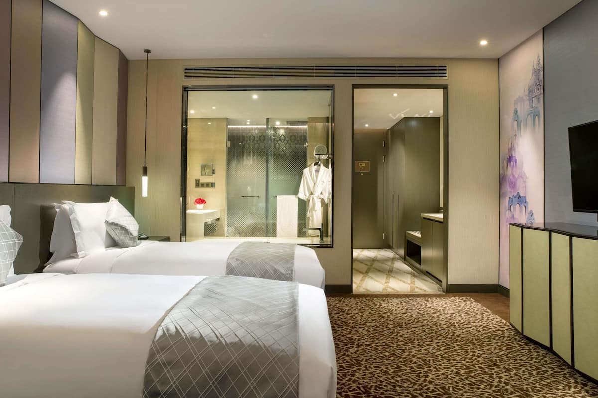 Habitación de hotel con dos camas, baño con ducha de vidrio y televisión sobre un mueble.