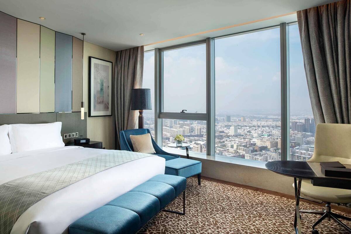 Habitación de hotel con una cama grande, un escritorio, una silla y vistas a la ciudad a través de amplias ventanas.