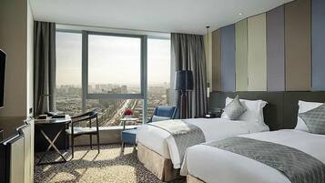 A hotel room with two beds, a desk, a chair, a TV, and a large window with a city view.