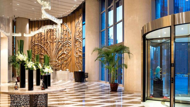 Een lobby met een marmeren vloer, een receptiebalie, een grote decoratieve wand en een glazen lift.