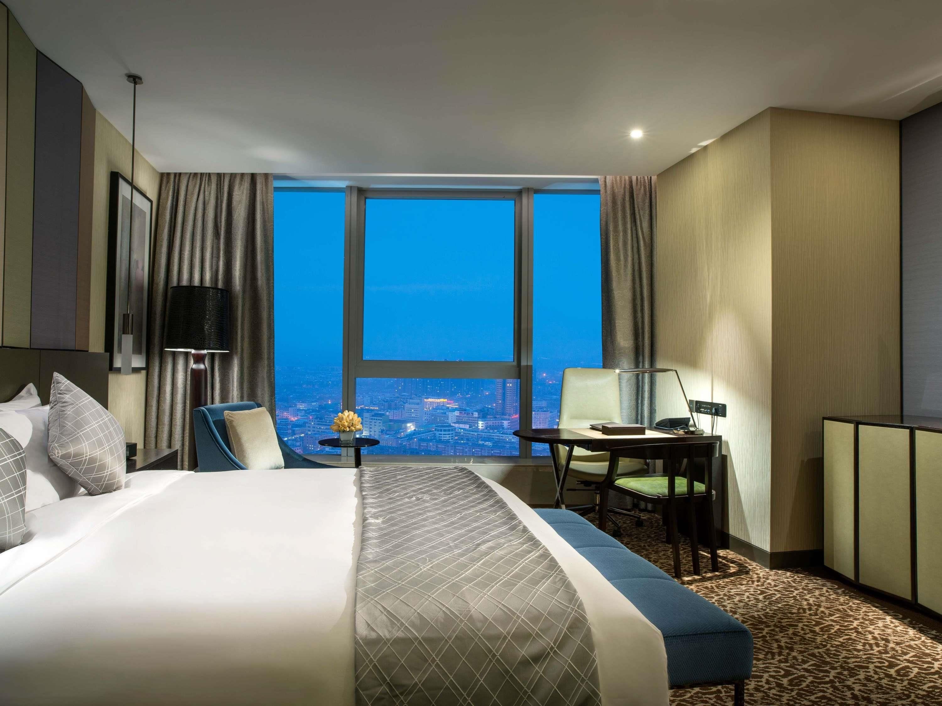 A hotel room with a large bed, a desk, a chair, and a view of the city at night.