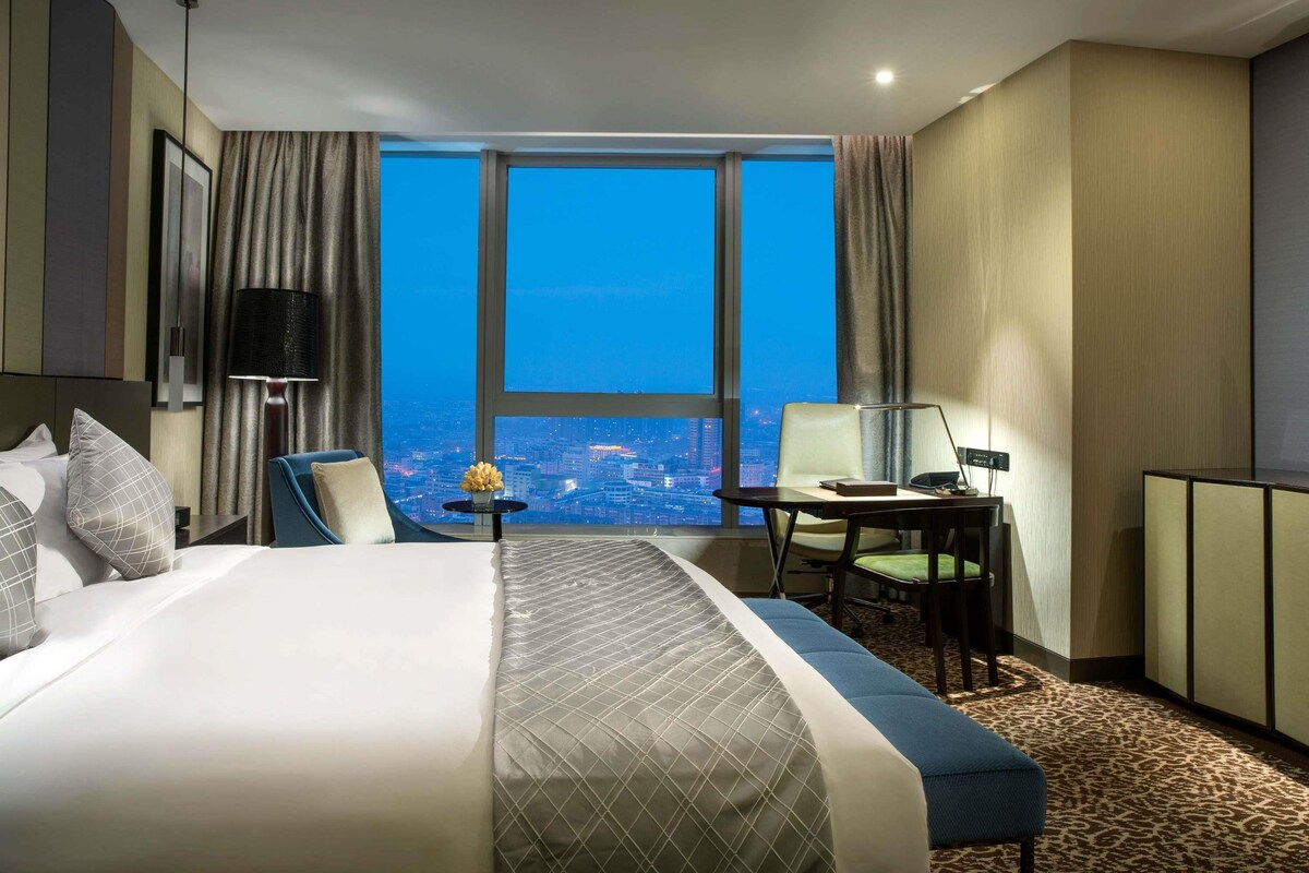 A hotel room with a large bed, a desk, a chair, and a view of the city at night.