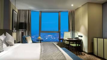 A hotel room with a large bed, a desk, a chair, and a view of the city at night.