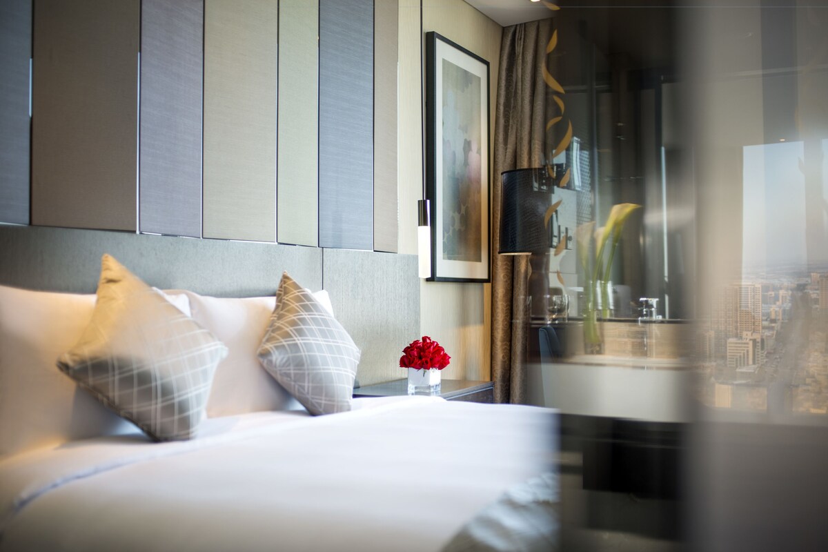 Een hotelkamer met een bed, kussens, een vaas met bloemen en uitzicht op een stadsbeeld door een raam.