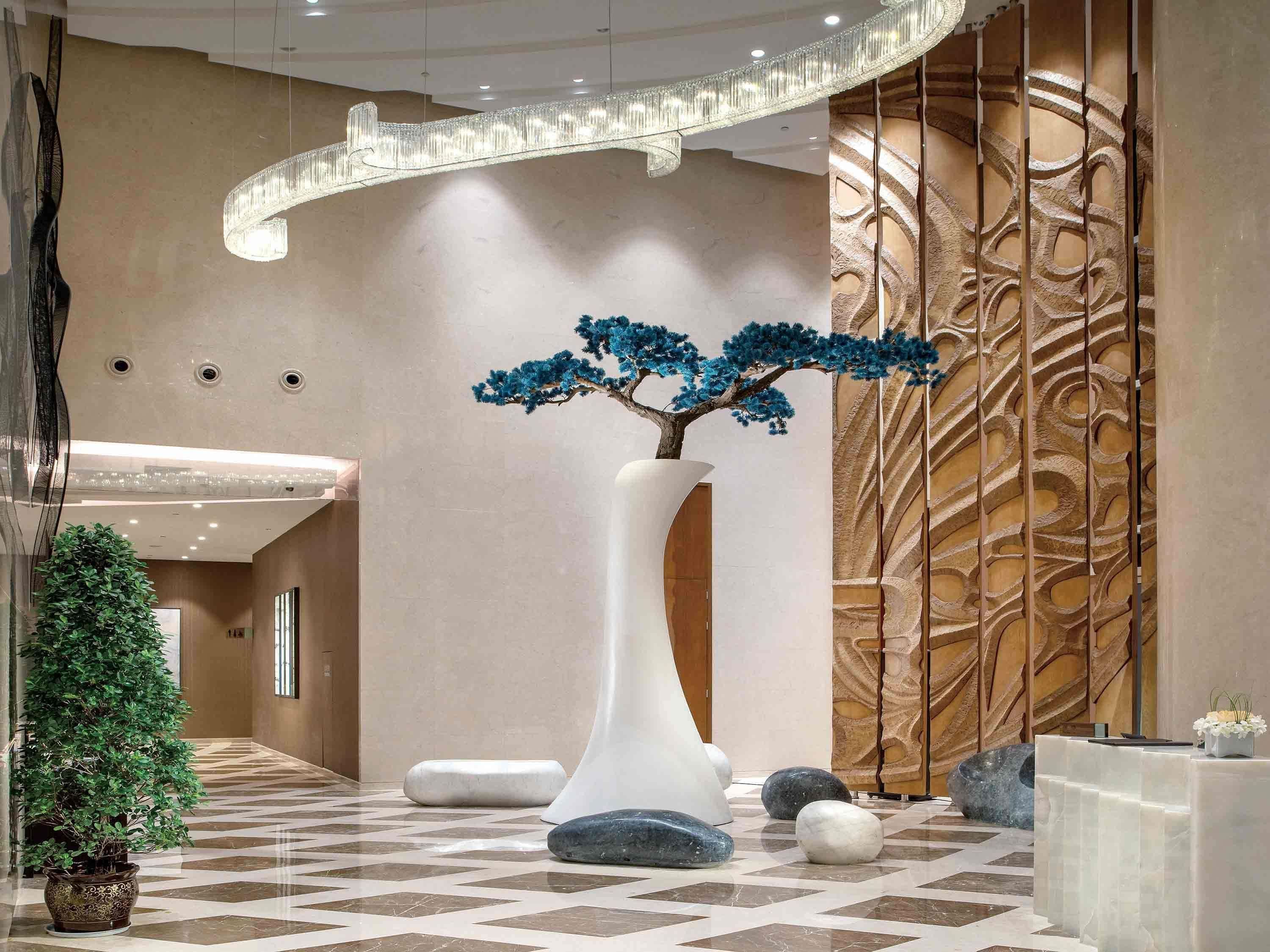 A lobby with a reception desk, a large white vase with a tree, a potted plant, and a patterned wall.