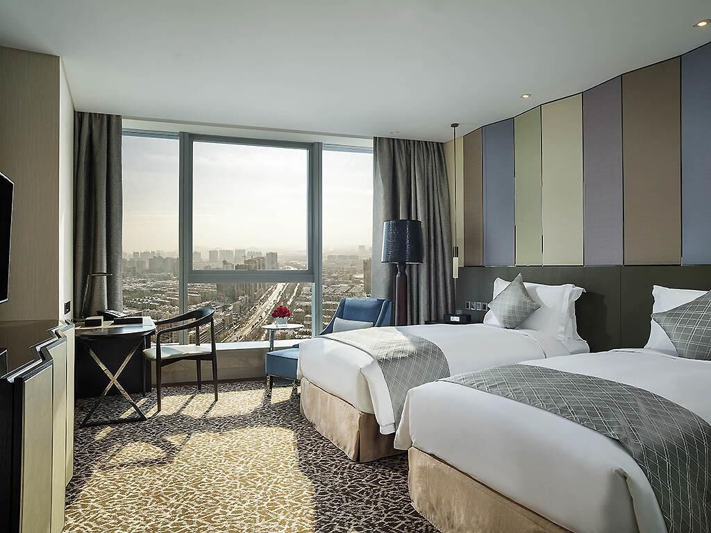 A hotel room with two beds, a desk, a chair, a TV, and a large window with a city view.