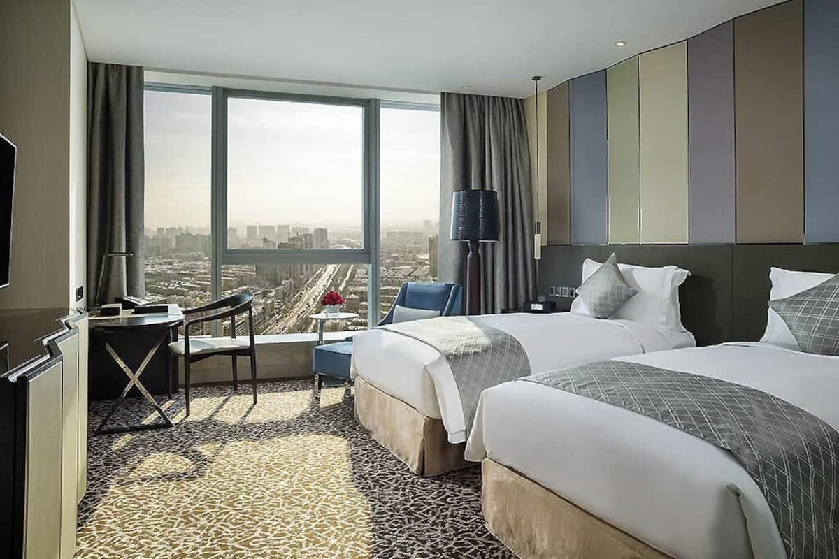 A hotel room with two beds, a desk, a chair, a TV, and a large window with a city view.