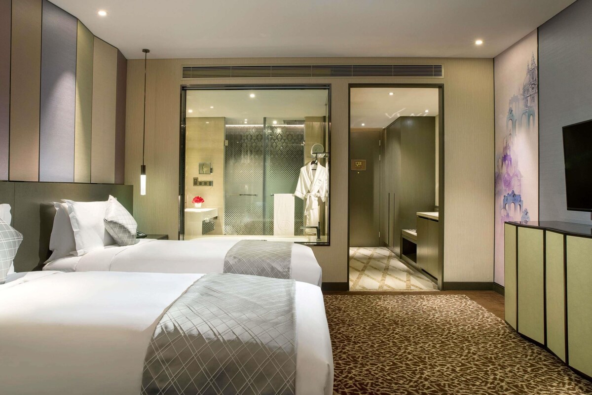 A hotel room with two beds, a bathroom with a glass shower, and a TV on a cabinet.