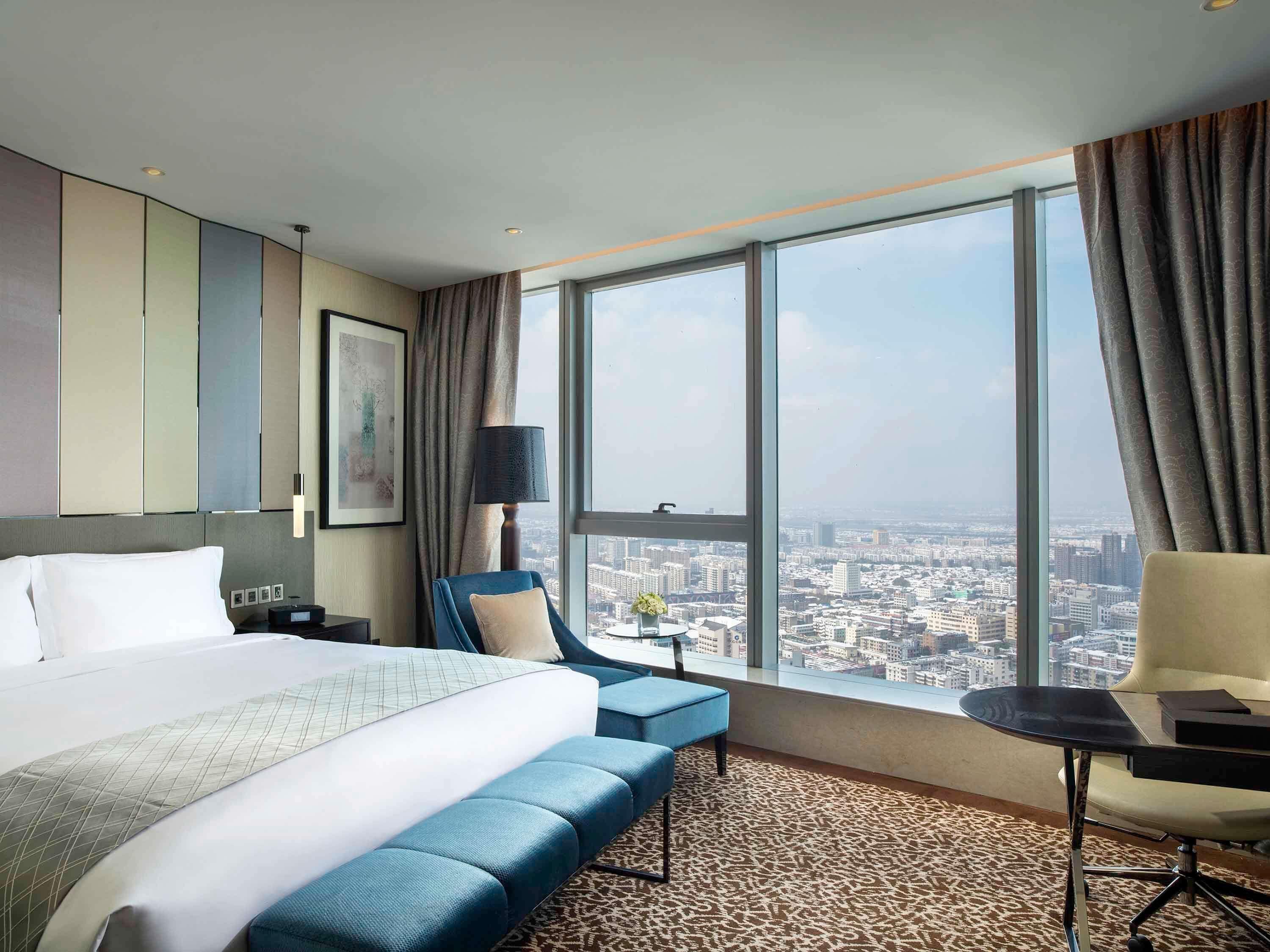 A hotel room with a large bed, a desk, a chair, and a view of a cityscape through large windows.