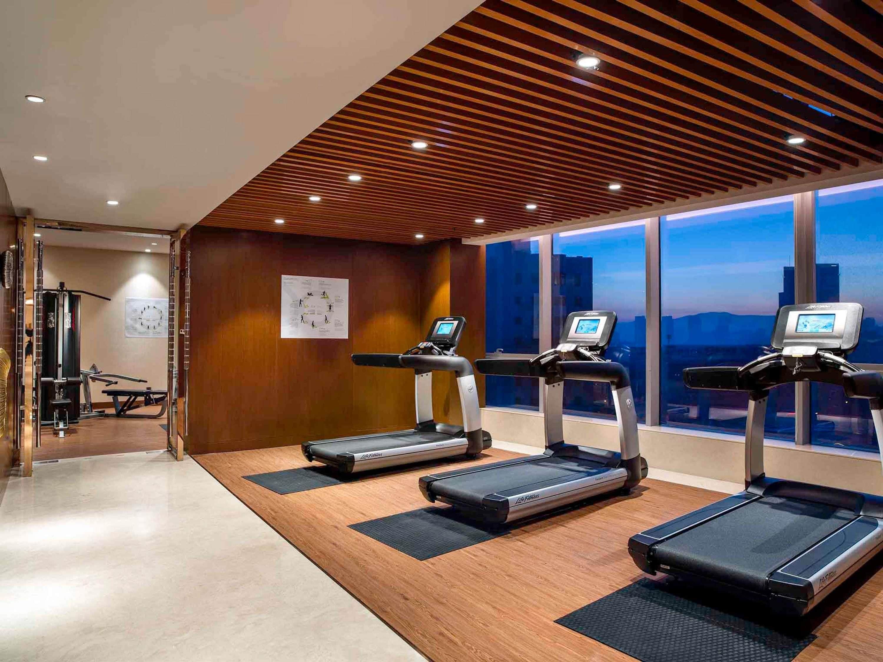Un gimnasio con cintas de correr, equipo de ejercicio y amplios ventanales con vistas a la ciudad.