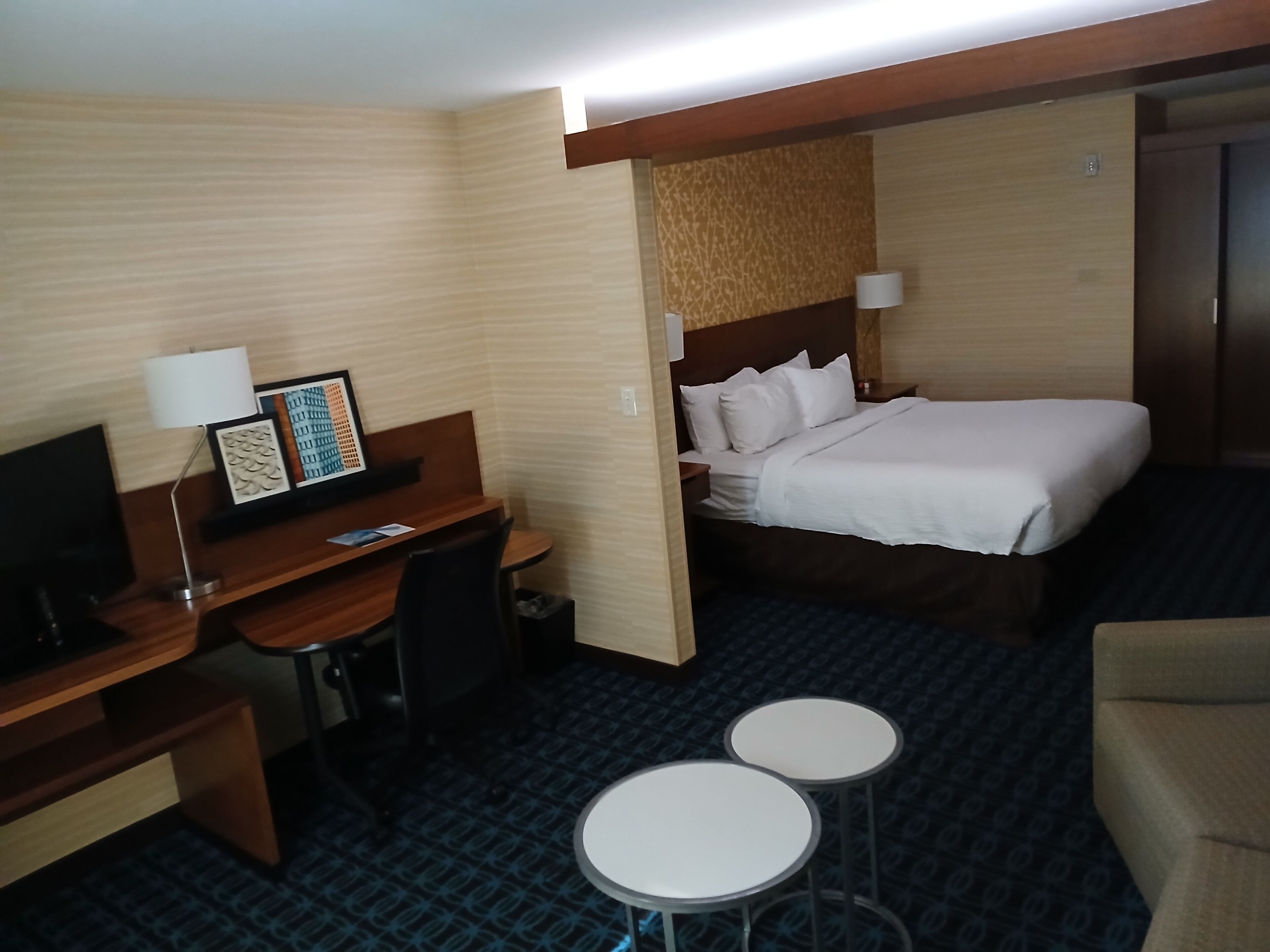 A hotel room with a bed, desk, chair, two small tables, and a TV.