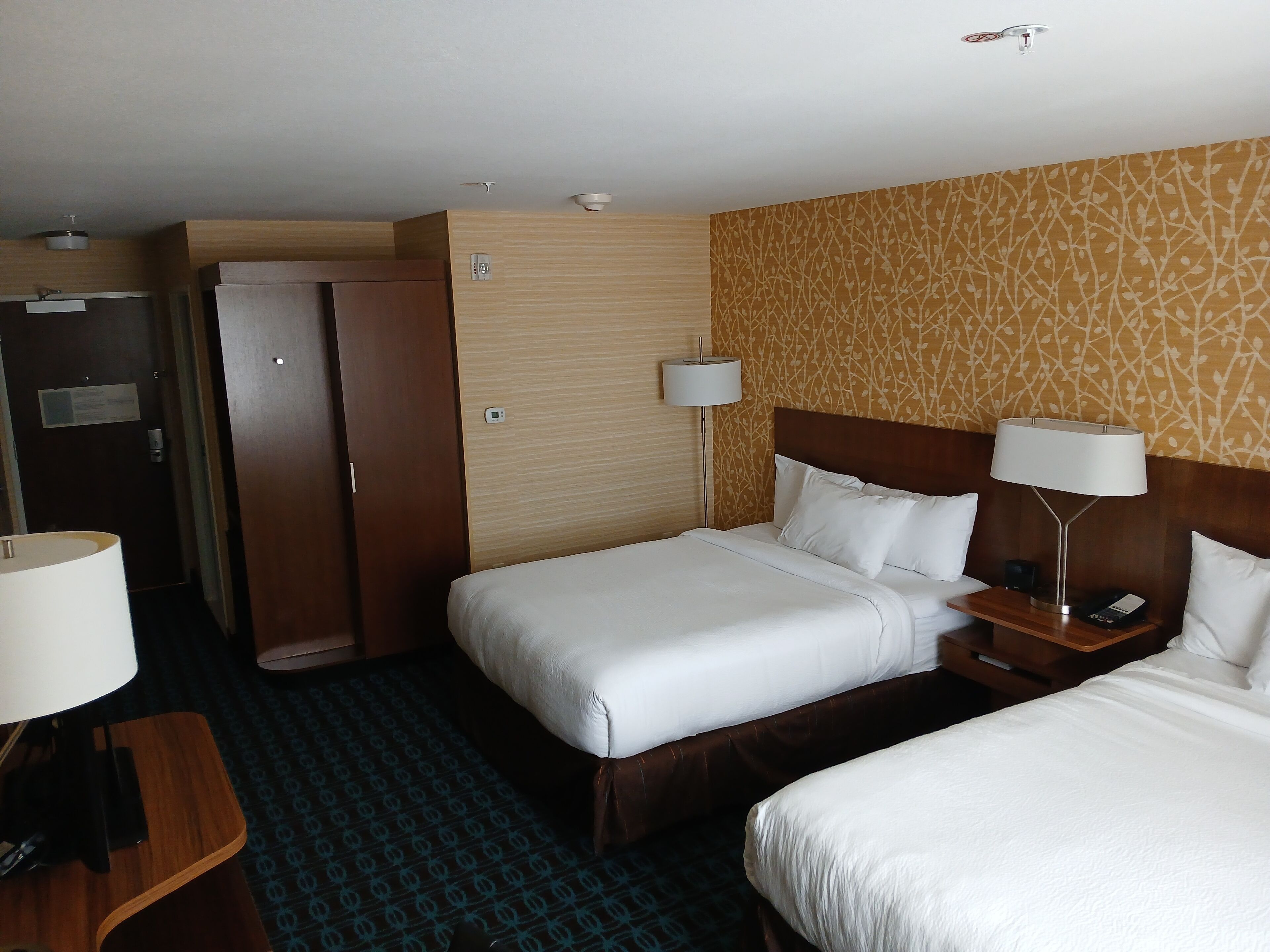 A hotel room with two beds, a wardrobe, a desk with a lamp, and a patterned wall.