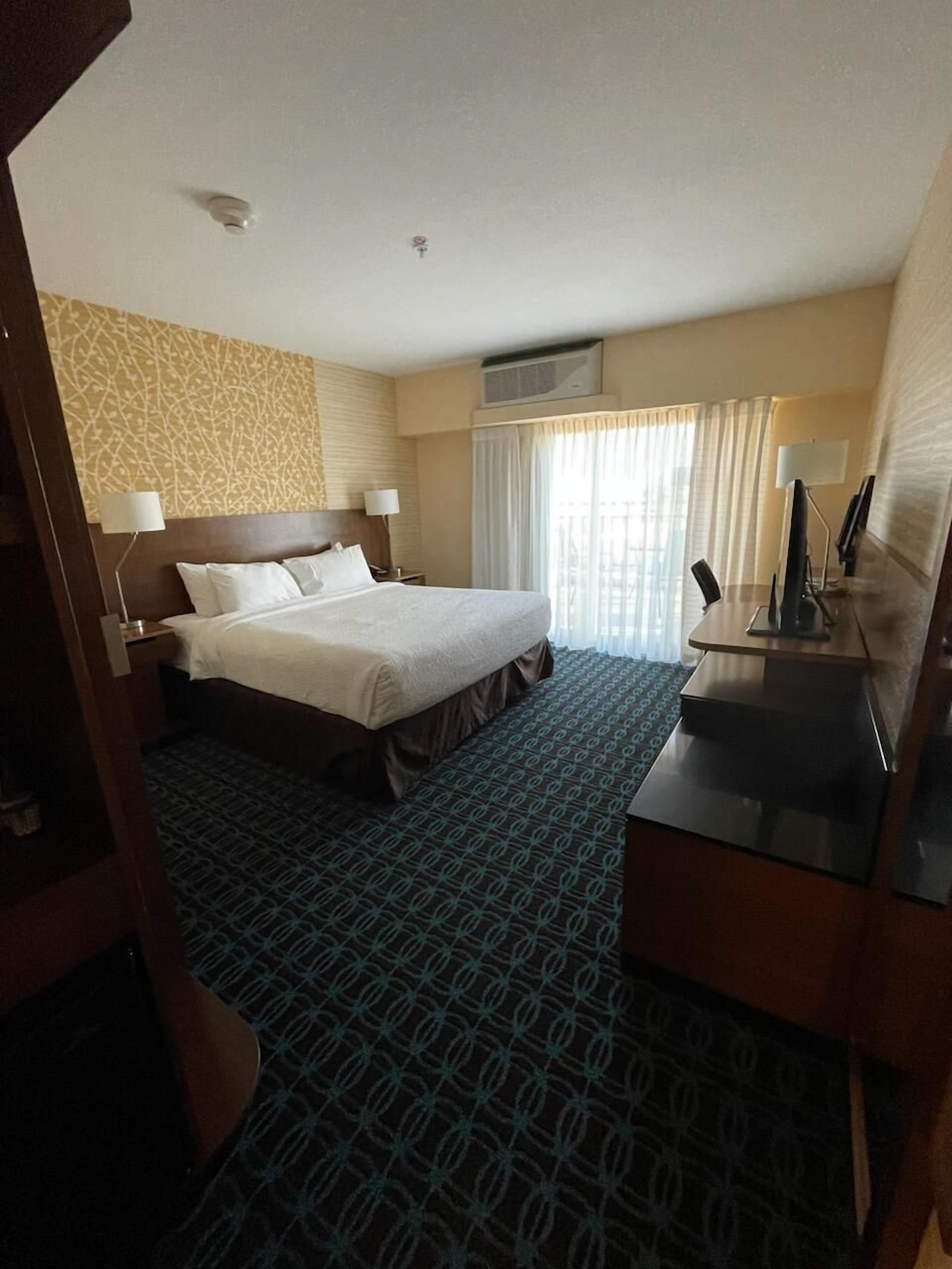 A hotel room with a large bed, a desk with a computer, a lamp, and a window with curtains.
