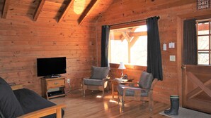 Interior de una cabaña de madera con televisión, dos sillones, una mesa y un ventanal con cortinas.
