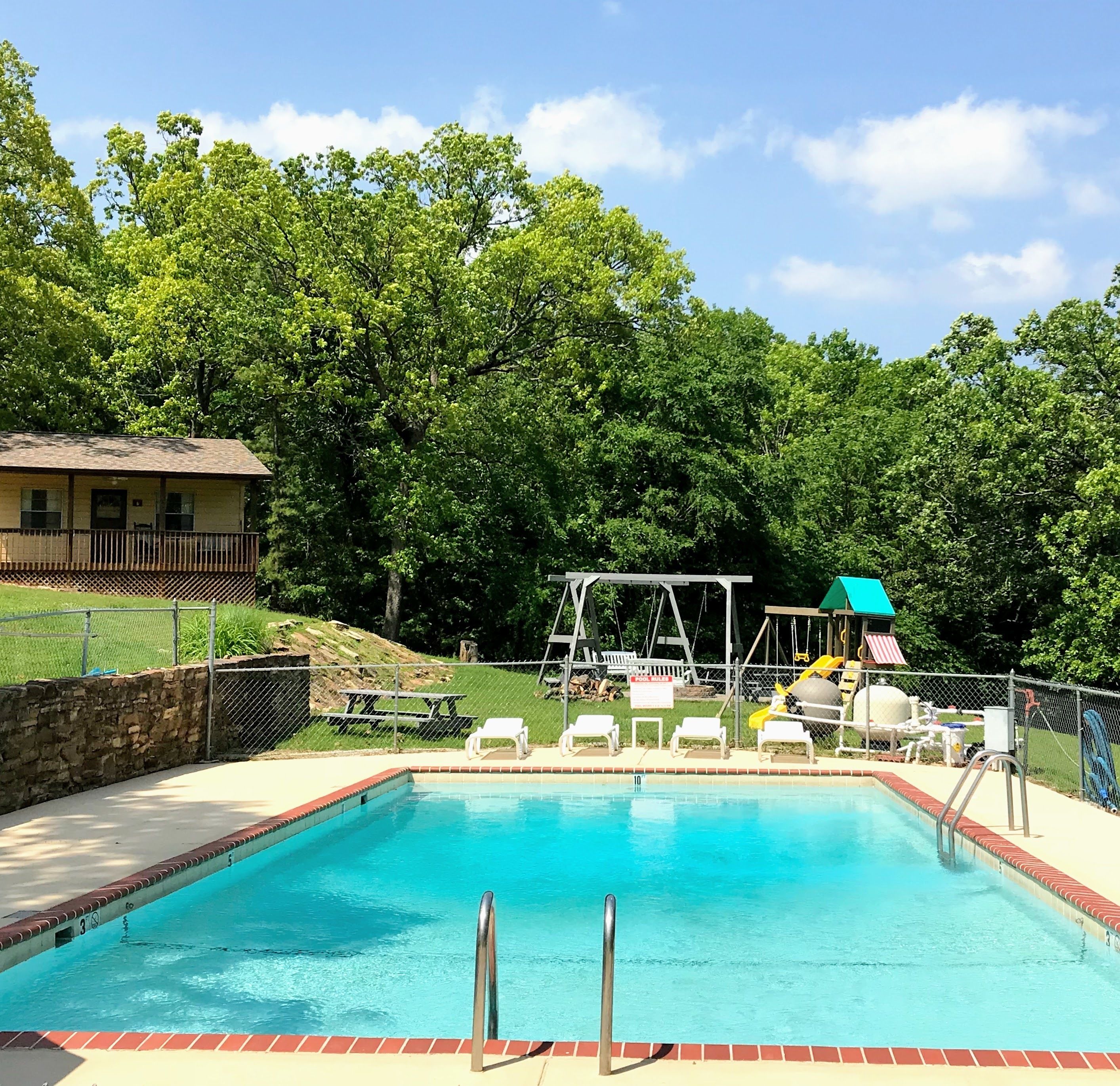 A rectangular swimming pool with a brick border, surrounded by a fence, and a picnic table area with chairs.