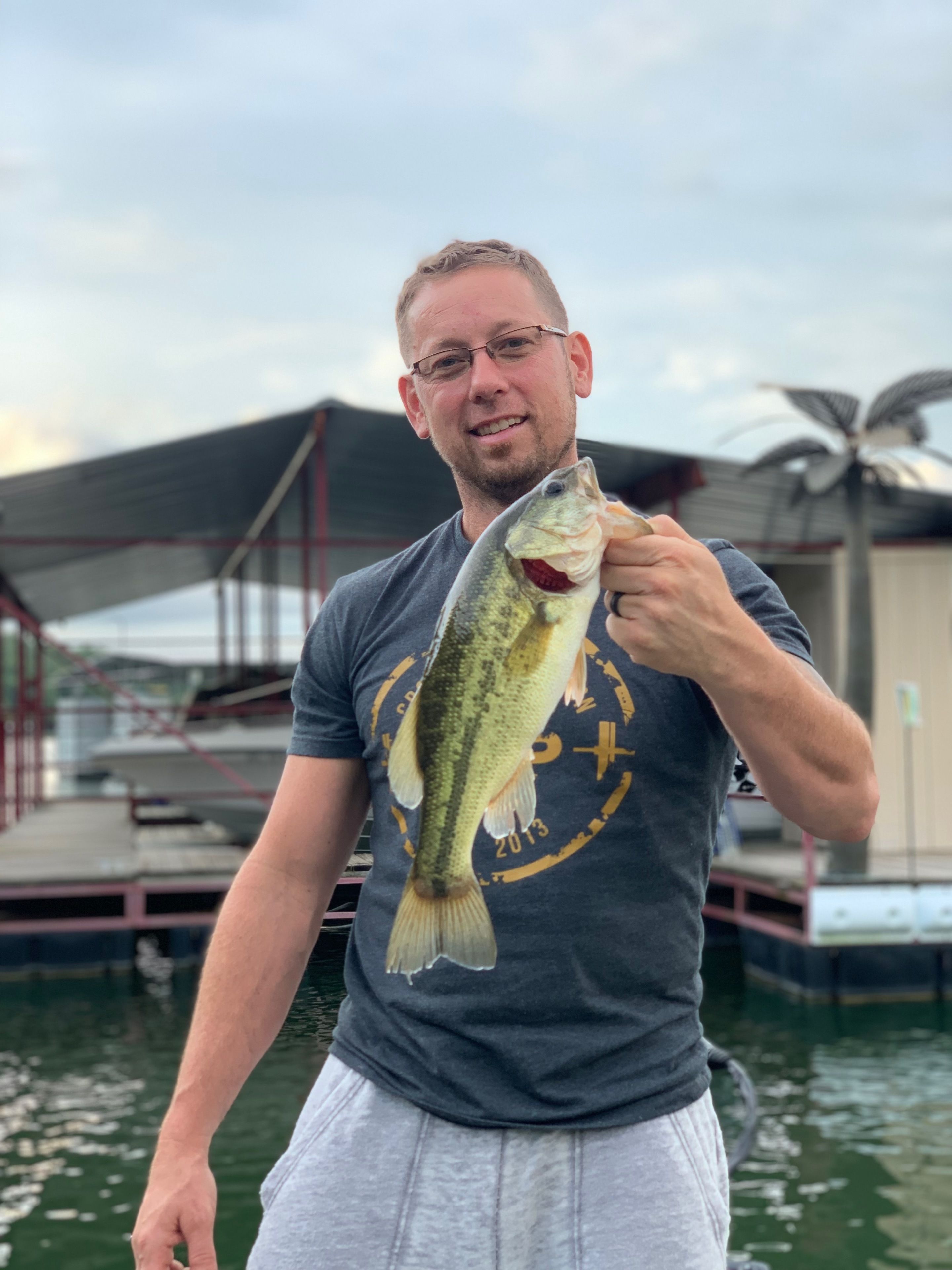 Una persona che tiene un pesce, indossa una t-shirt con un logo e si trova vicino a una distesa d'acqua con una struttura sullo sfondo.