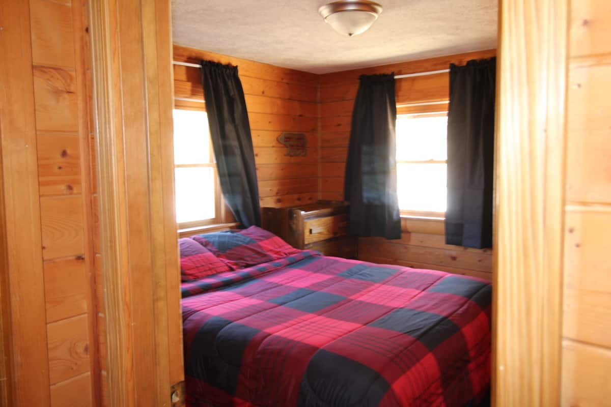 Une chambre dans une cabane en bois, avec un lit, deux fenêtres avec des rideaux et un luminaire au plafond.