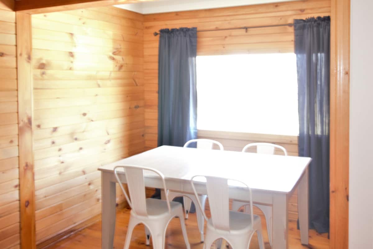 Une pièce aux murs lambrissés, avec une table blanche et quatre chaises blanches, une grande fenêtre ornée de rideaux bleus et un plafond en bois.