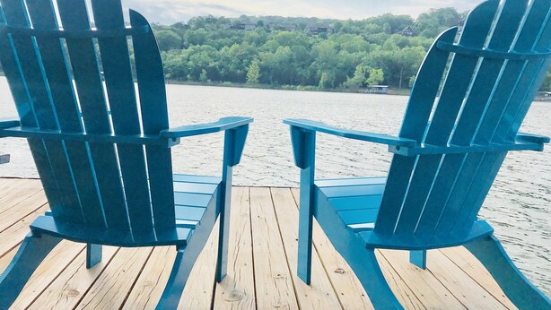 Dos sillas Adirondack azules en una terraza de madera con vistas a un lago y una orilla boscosa.