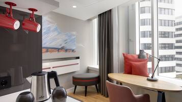 Una habitación de hotel moderna con una zona de cocina, una mesa de comedor con una silla, una ventana con vistas a edificios y un cuadro en la pared.