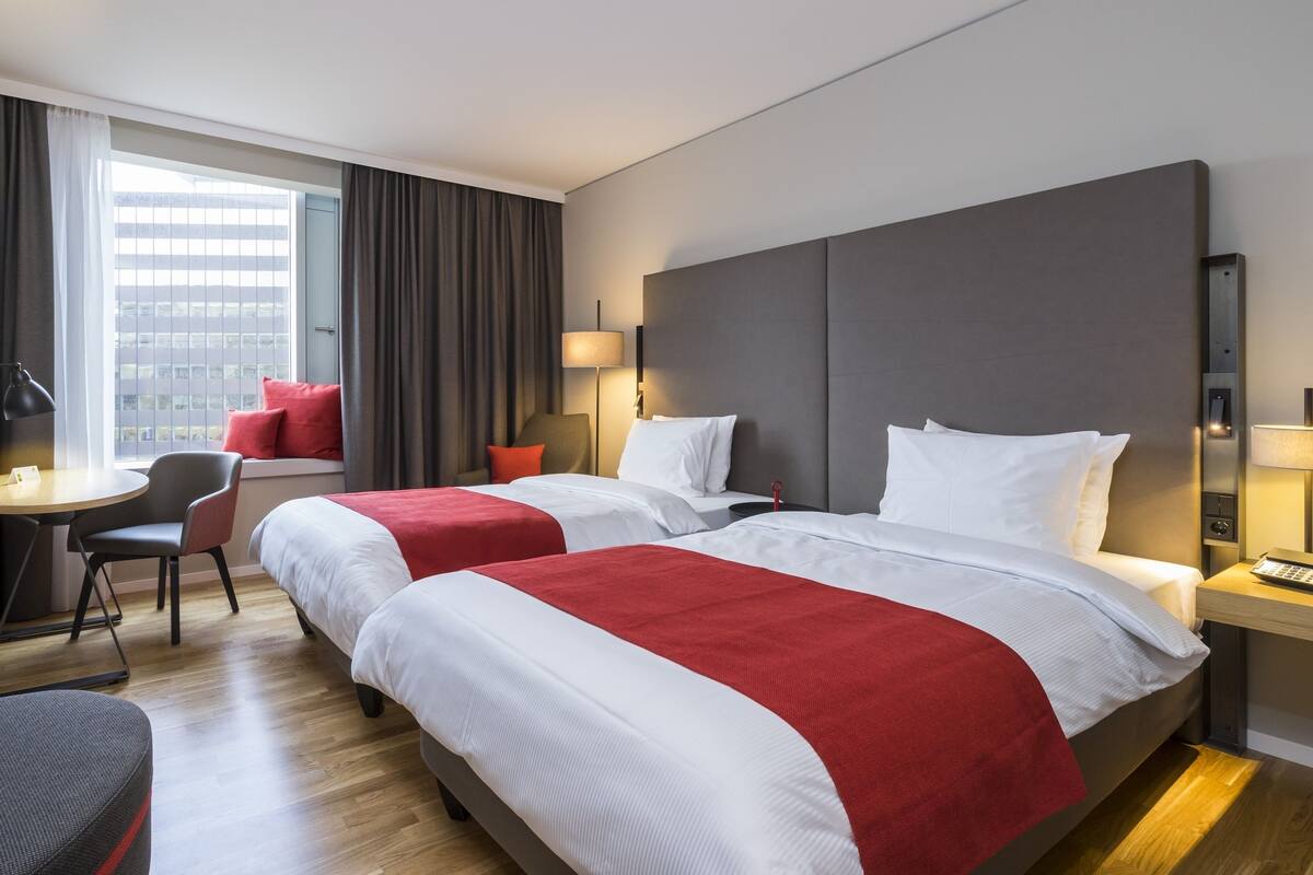 Une chambre d’hôtel avec deux lits, un bureau, une chaise, une télévision et une fenêtre avec des rideaux.