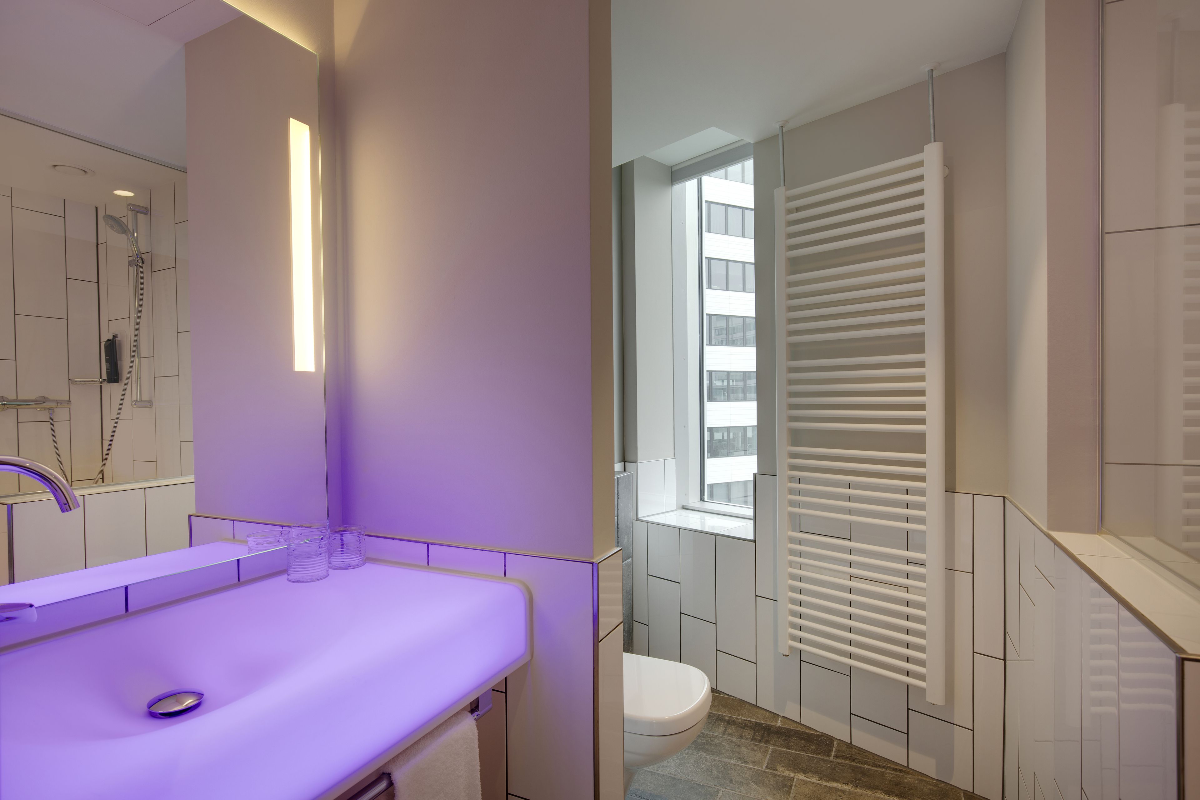 A modern bathroom with a purple-lit vanity, a shower, and a toilet with a window and blinds.
