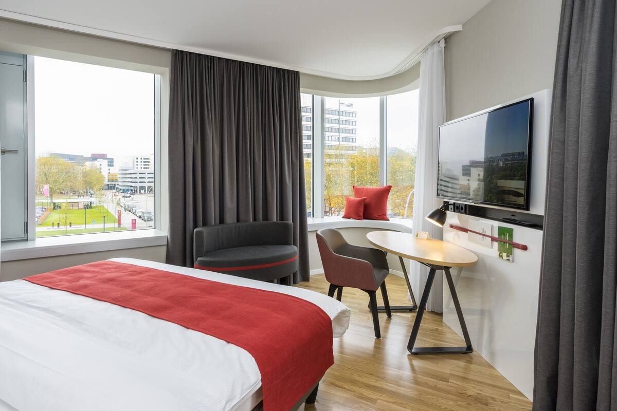 Une chambre d’hôtel équipée d’un lit, d’un bureau, d’une chaise, d’une télévision et de grandes fenêtres donnant sur la ville.