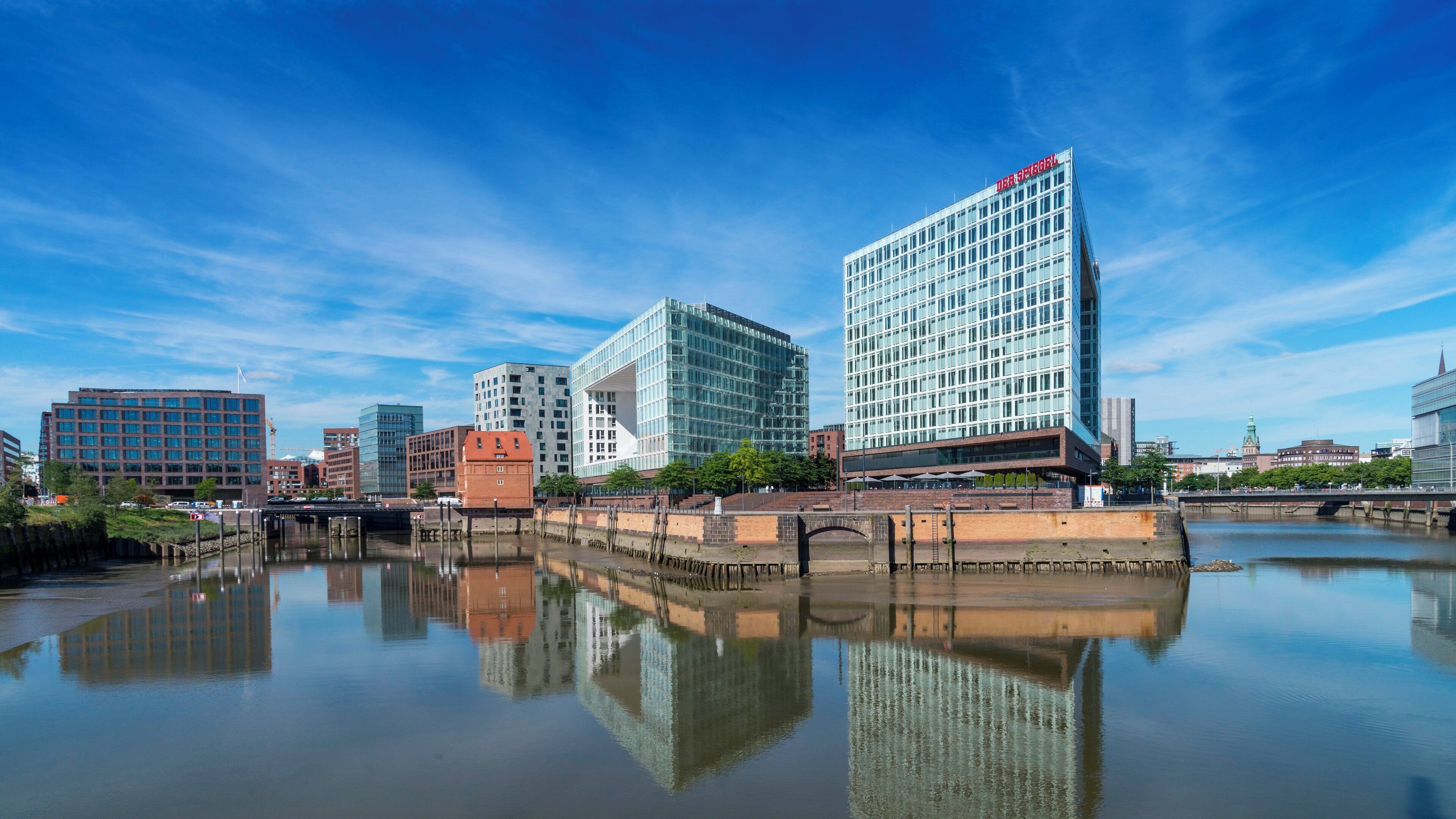 A cityscape with modern buildings, a calm river reflecting the structures, and a clear blue sky.