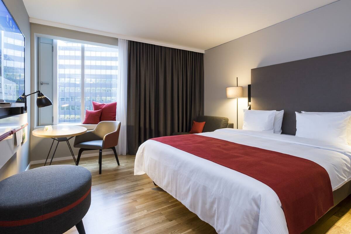 Une chambre d’hôtel avec un grand lit, un bureau, une chaise, une petite table et une vue sur la ville par la fenêtre.