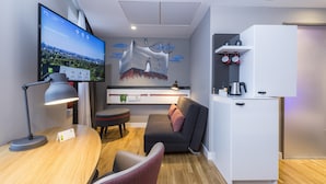 Ein modernes Hotelzimmer mit einem Flachbildfernseher, der eine Stadtansicht zeigt, einem Holztisch mit Lampe, einem grauen Sofa mit einem rosafarbenen Kissen, einem weißen Schrank mit Wasserkocher und einem Wandregal mit Tassen.